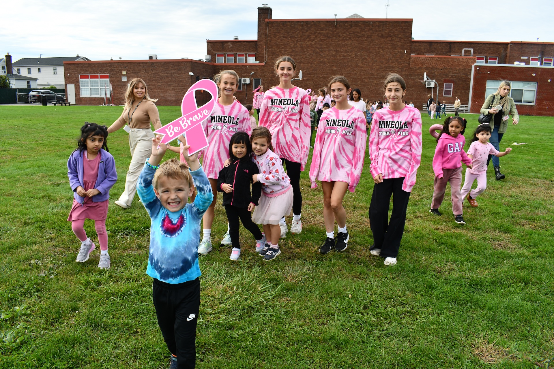 a young student holds up a pink ribbon while leading a parade including high school students on a grass field.